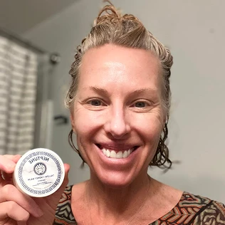 A smiling woman with blonde hair holds up a small, round, white container.