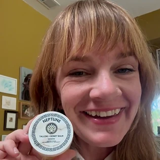 A smiling woman with bangs holds up a small, round white container of Neptune Tallow + Honey Balm.