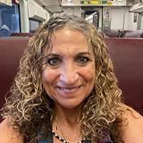 A woman with long, curly, highlighted hair smiles at the camera while sitting on a train or bus.