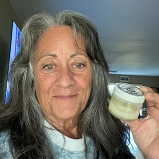 A woman with long, graying hair holds up a small, white jar of a cream-like substance.