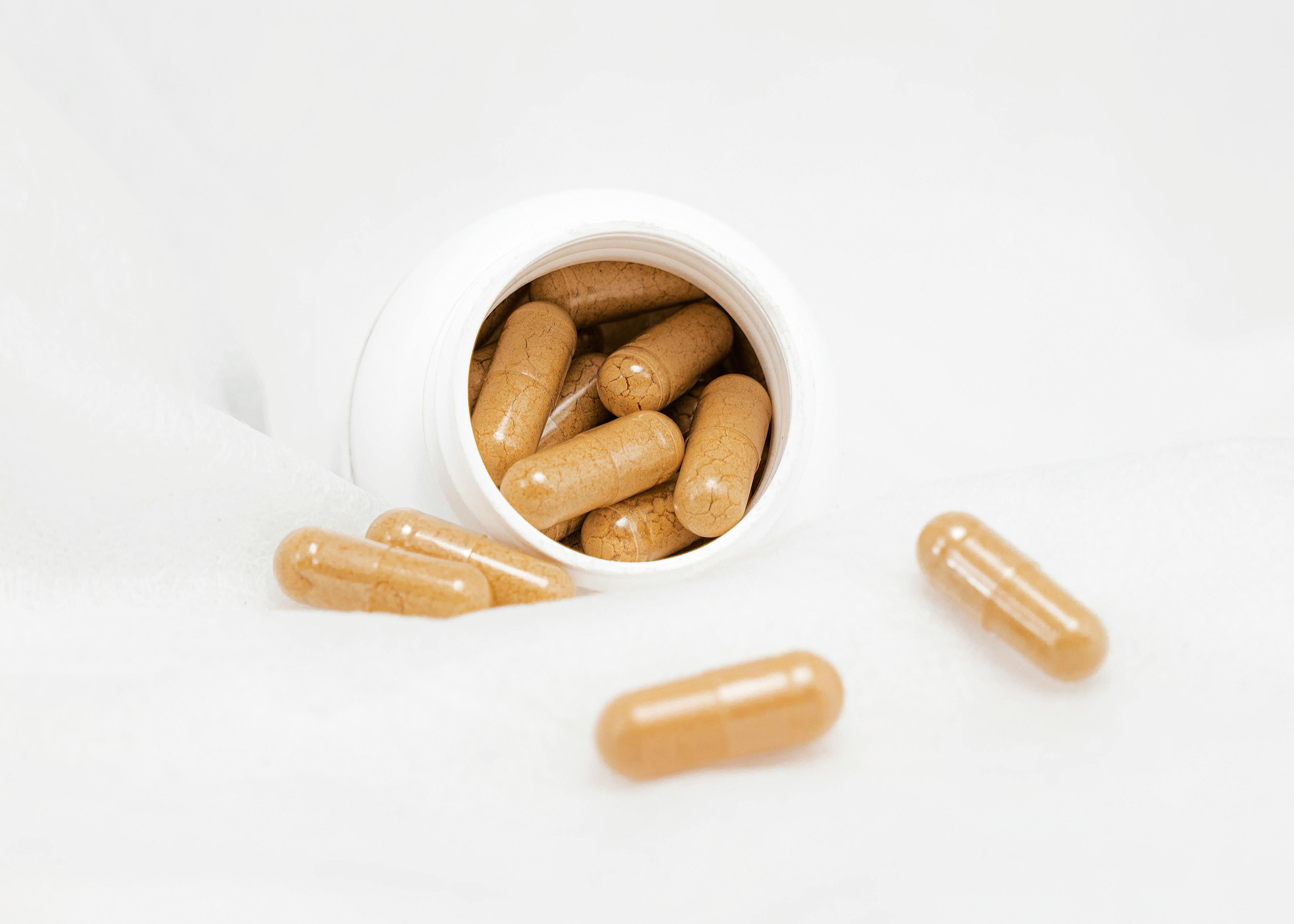 A close-up of a white bottle tipped over, with brownish capsules spilling out onto a white surface.