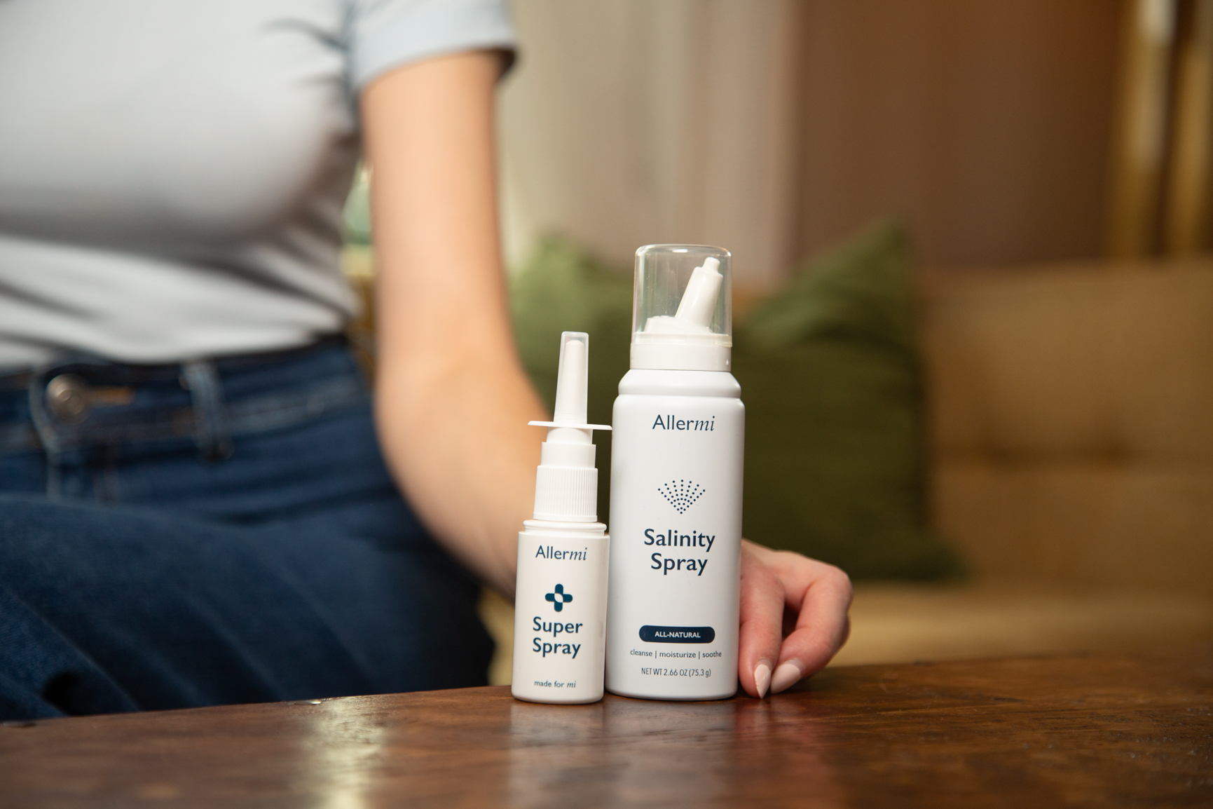 A person sits with two Allermi nasal spray products, a Super Spray and a Salinity Spray, on a table.