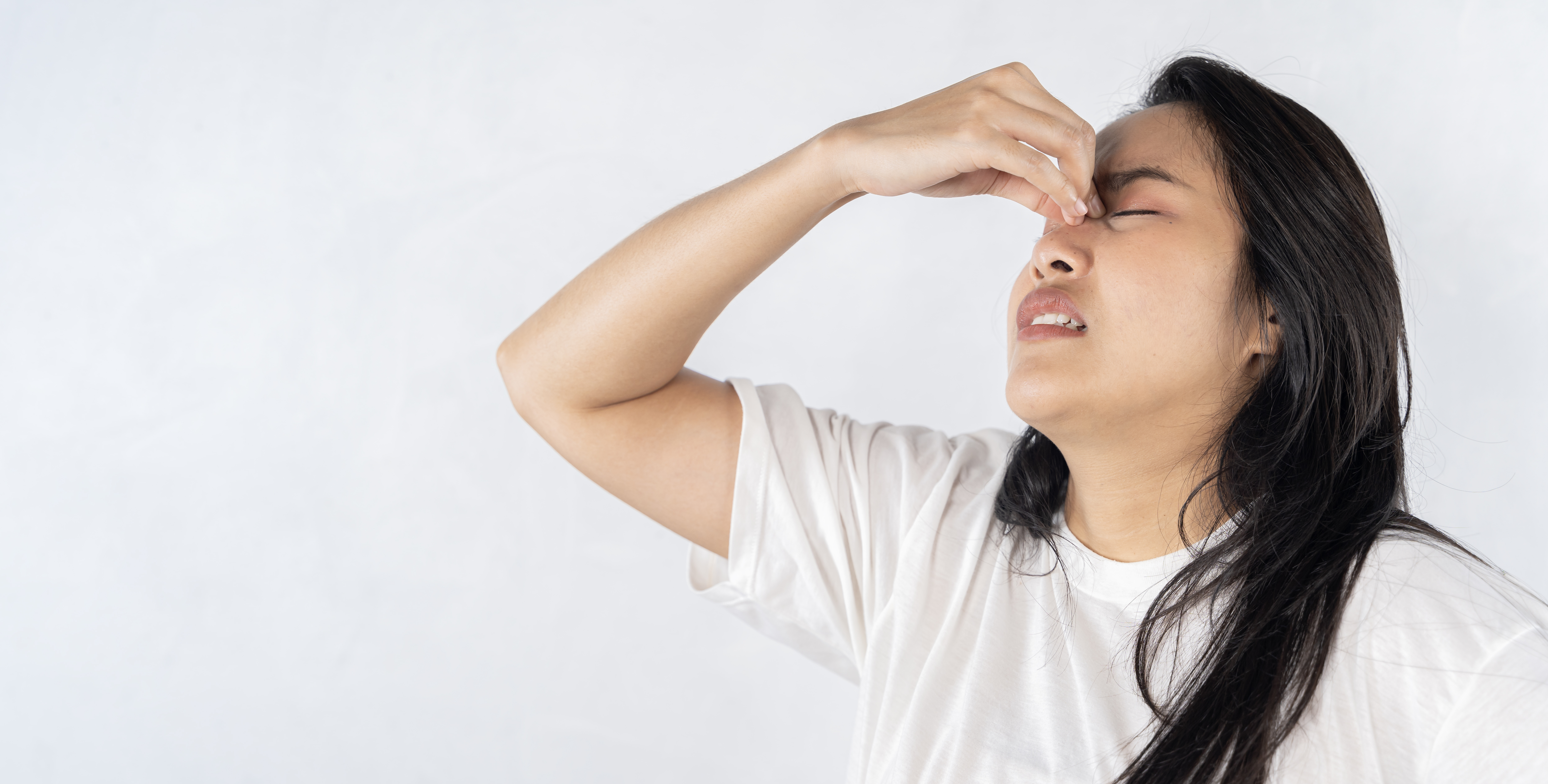A woman in a white t-shirt pinches the bridge of her nose with a pained expression.