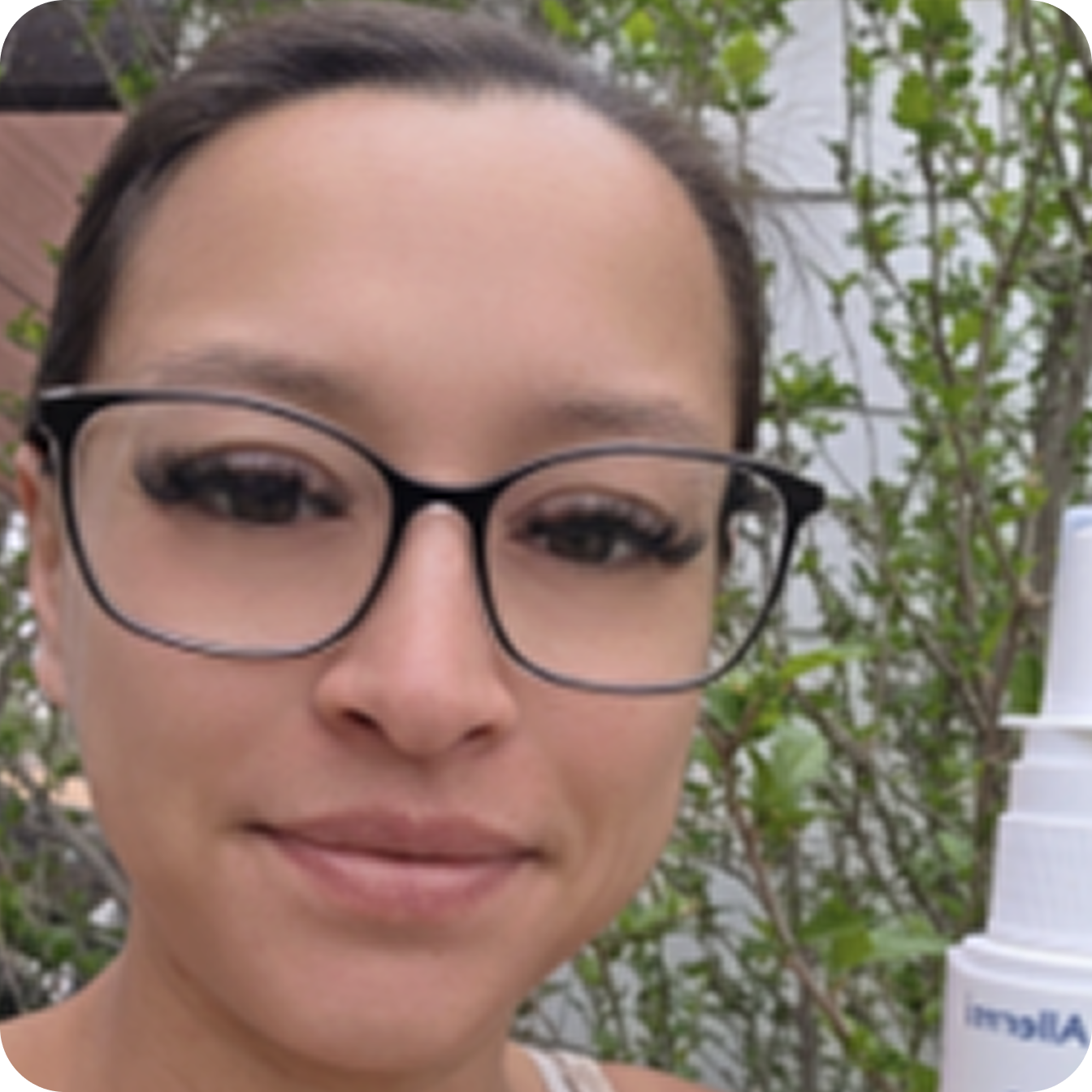 A close-up of a smiling woman with glasses holding a white bottle outdoors.