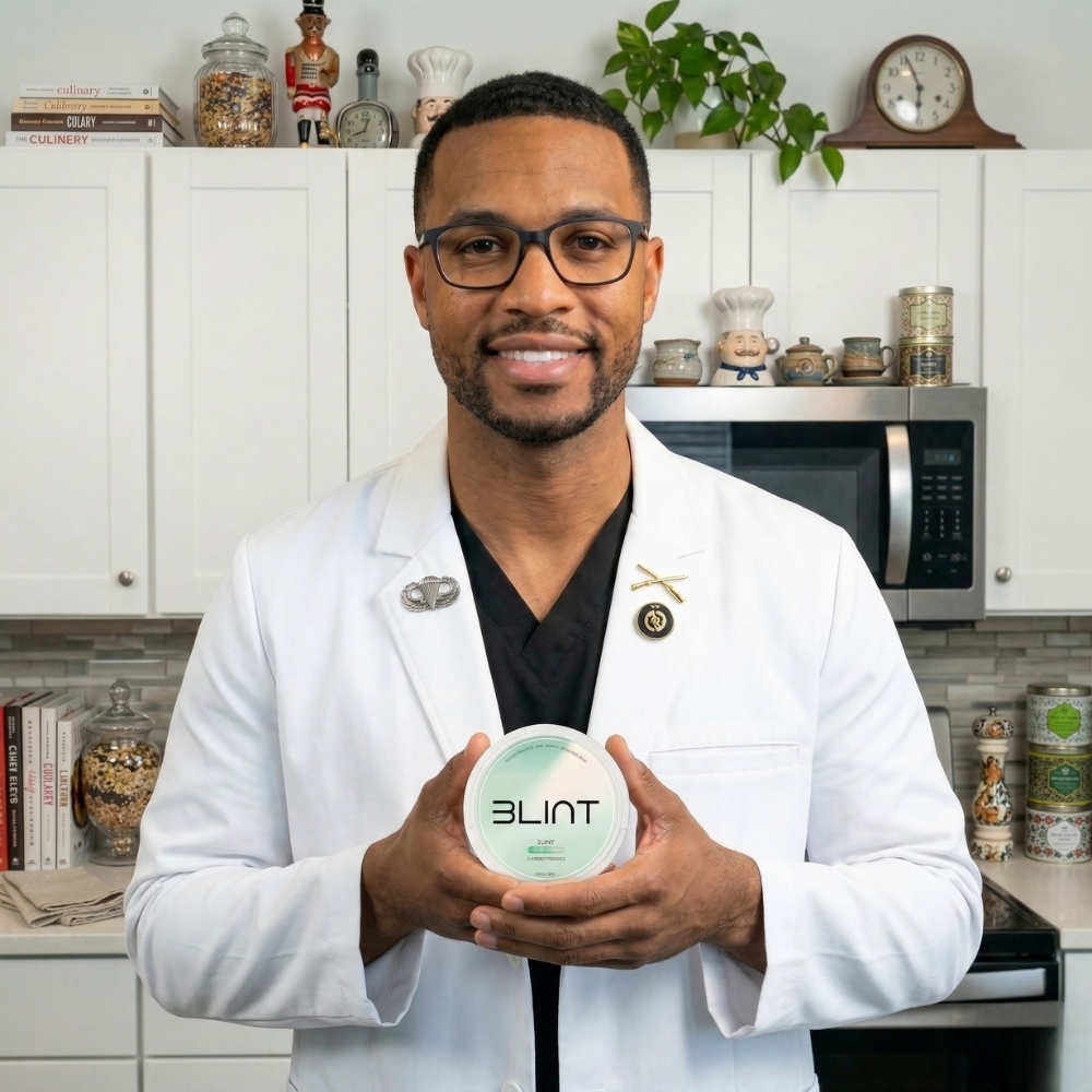 A smiling man in a white lab coat and glasses holds a round white container in a kitchen.