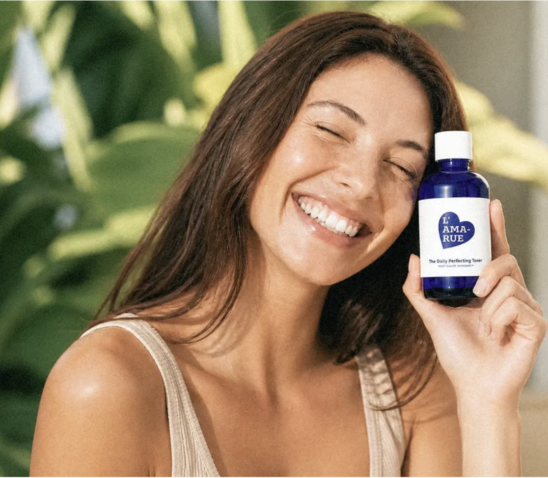 A woman with her eyes closed smiles while holding a blue bottle of L'AMARUE The Daily Perfecting Toner.