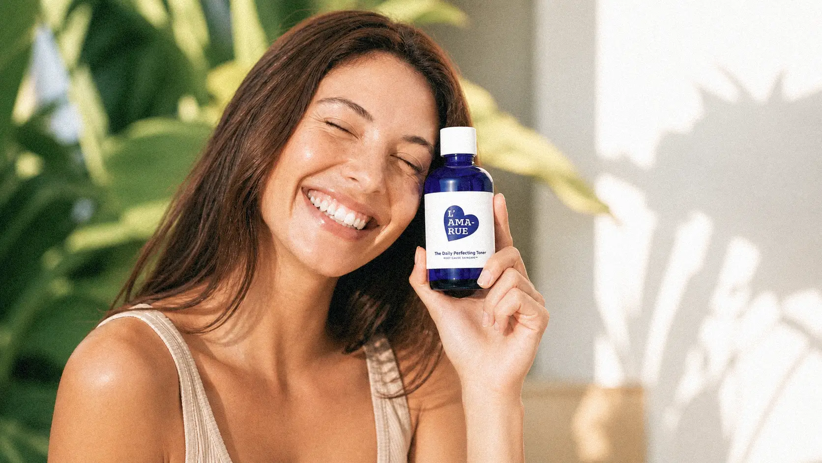 A smiling woman with her eyes closed holds a blue bottle of L'AMA-RUE The Daily Perfecting Toner.