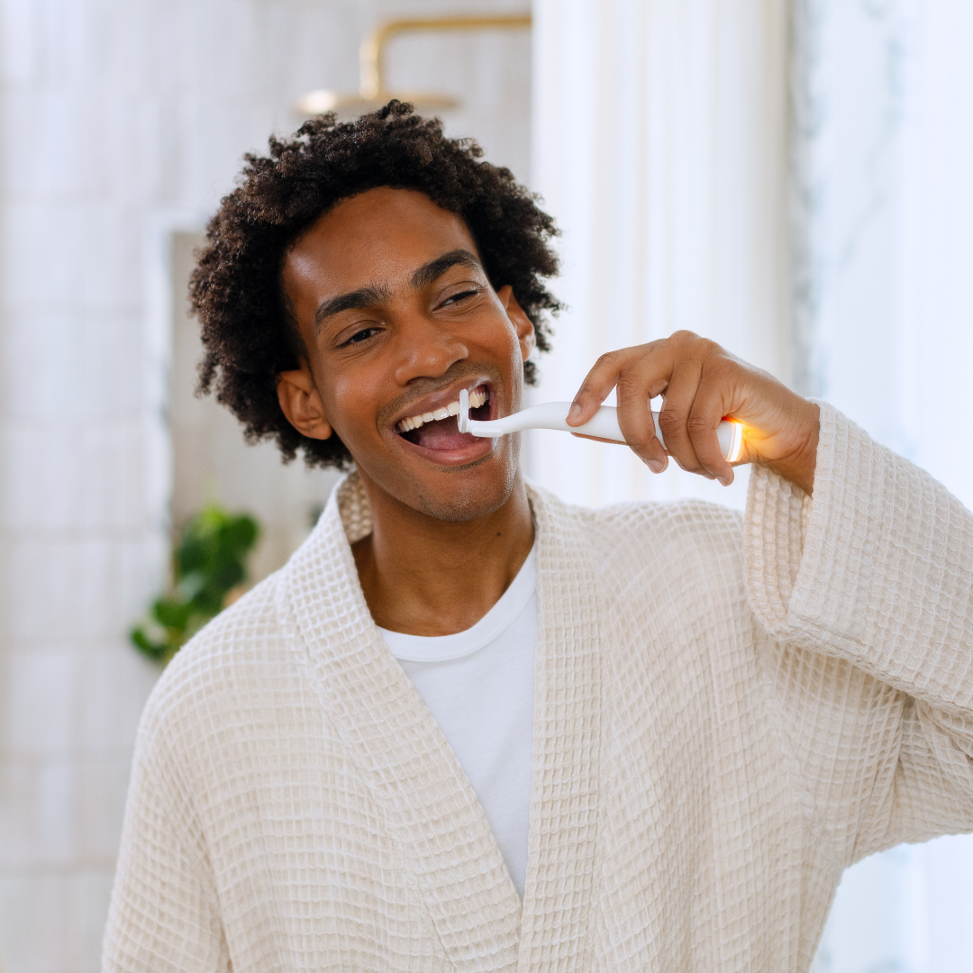 A smiling man in a waffle-knit robe brushes his teeth with a white electric toothbrush in a bathroom.
