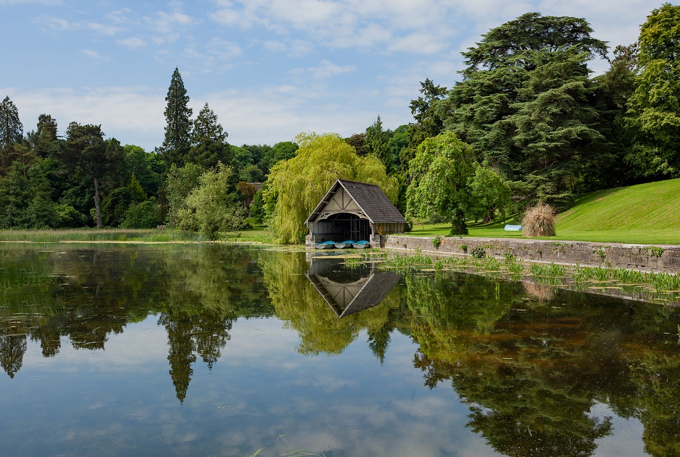 A boathouse by a lake surrounded by lush green trees and clear sky reflection.