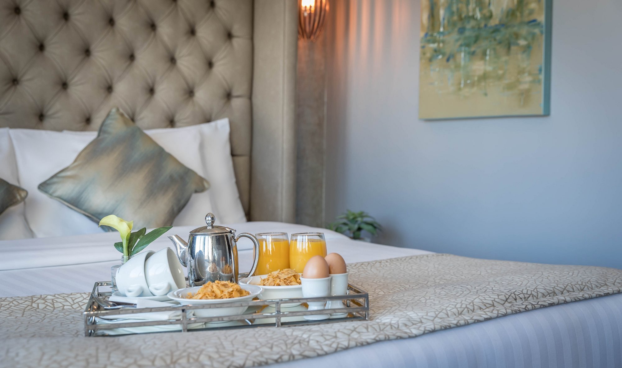 Breakfast tray on a bed with pillows and a painting on the wall.