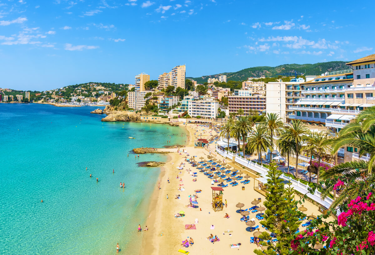 Sunny beach with turquoise water, people sunbathing, and buildings in the background.