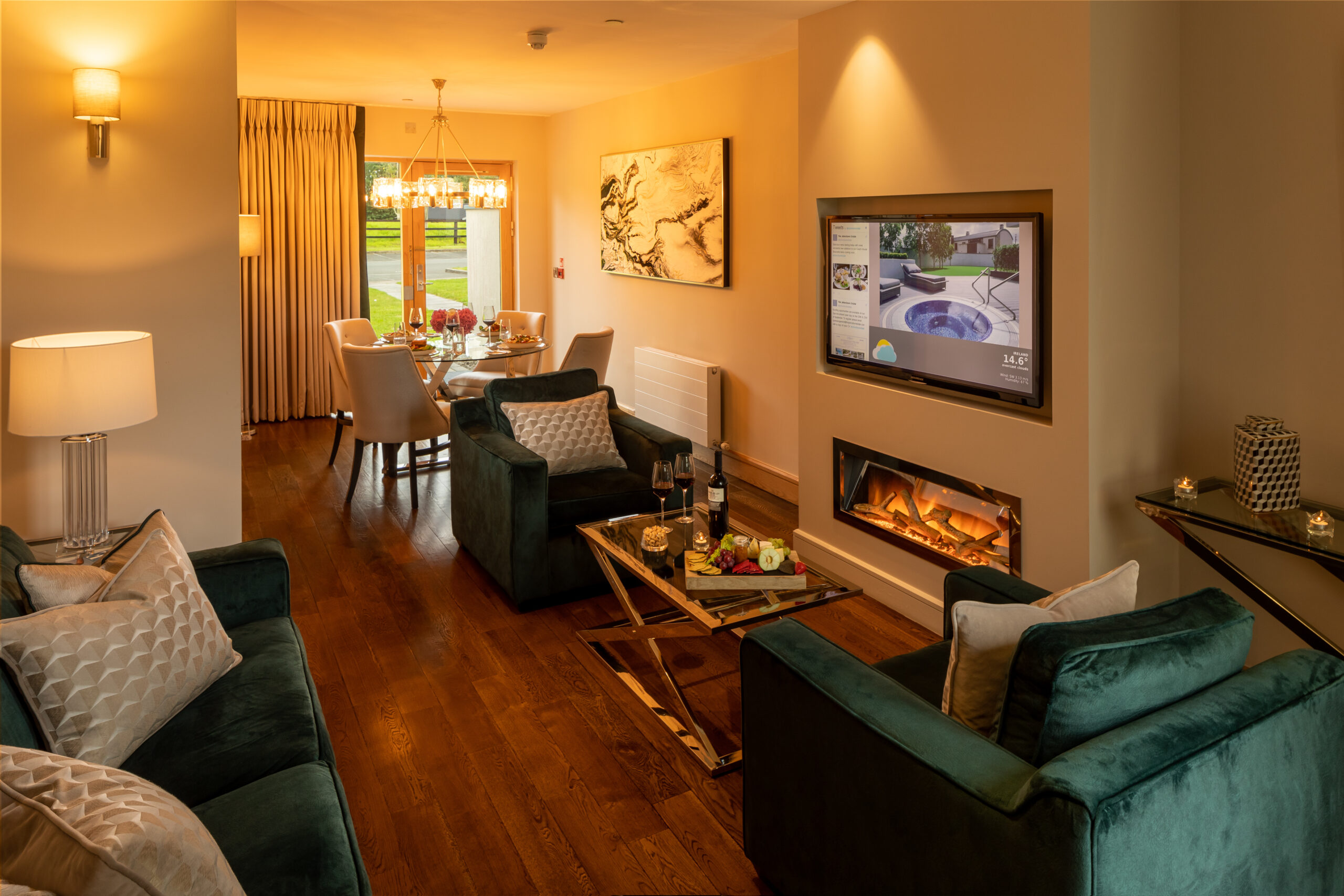 A warmly lit, luxurious living room with green velvet furniture, a fireplace, and a dining area in the background.