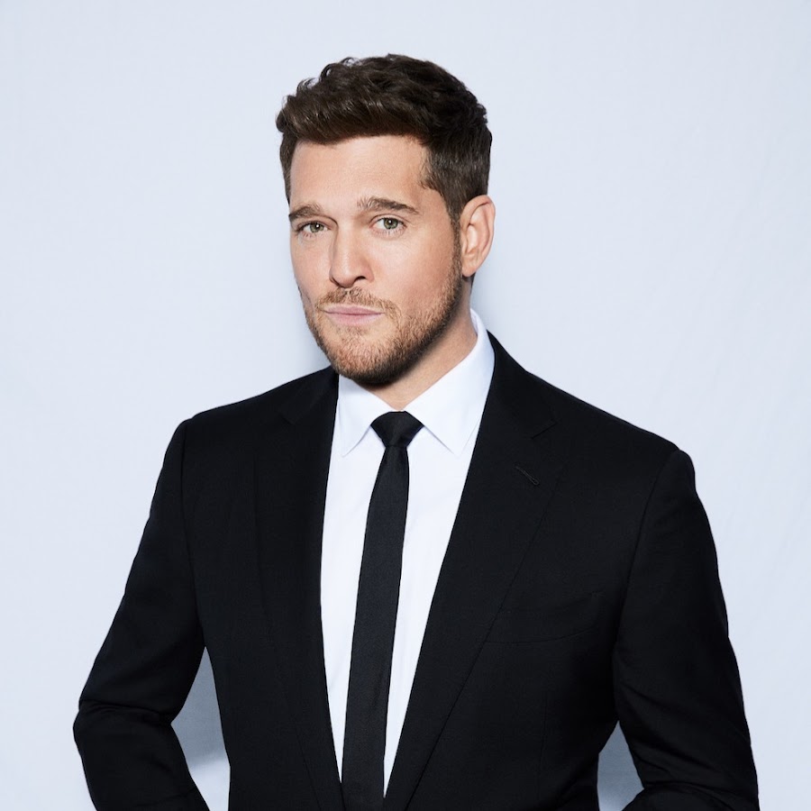 A man with brown hair and stubble wearing a black suit, white shirt, and black tie.