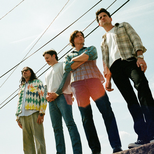 A low-angle photo of four men standing outdoors under a bright sky with power lines overhead.