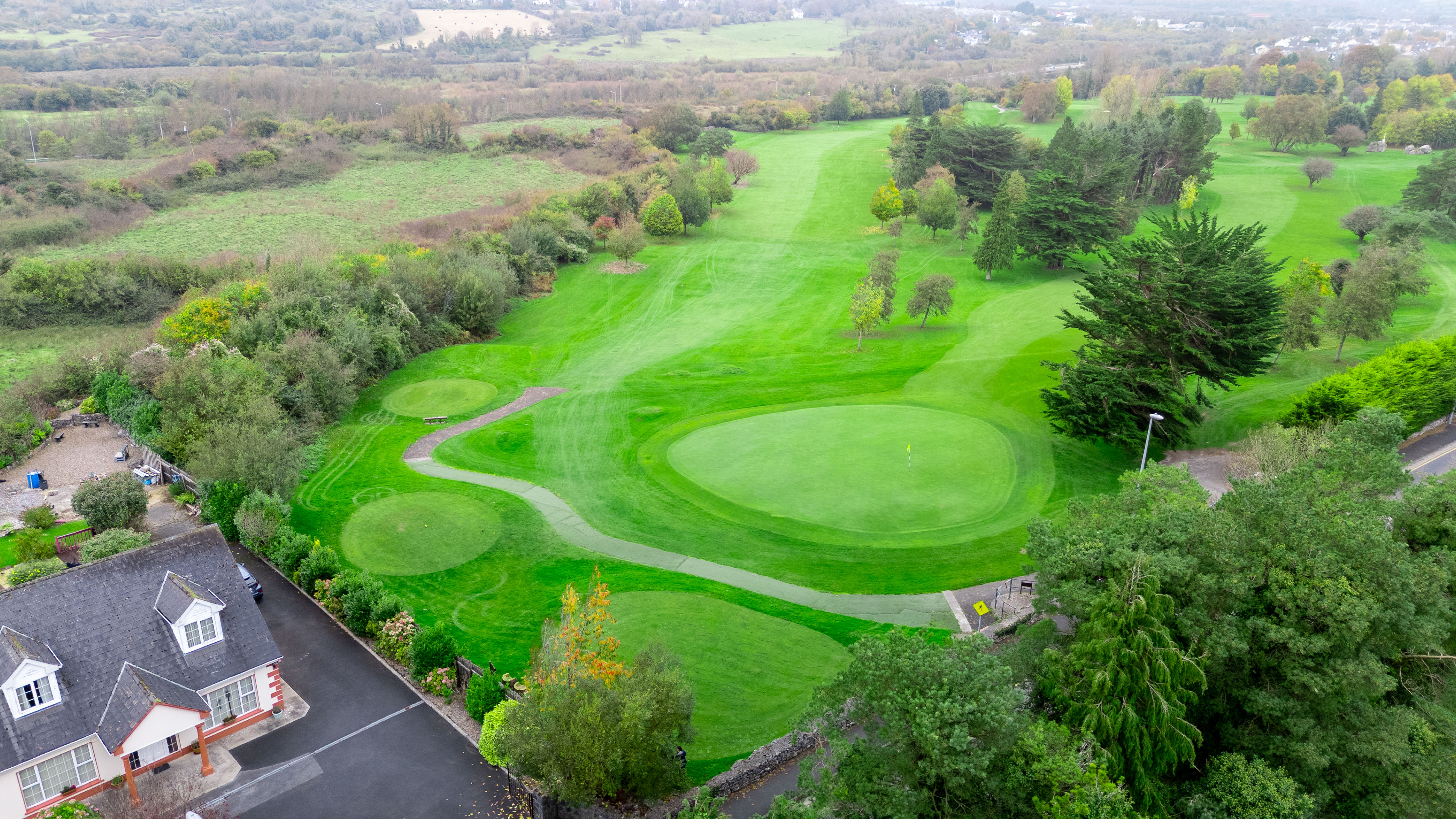 An aerial view of a lush, green golf course surrounded by trees and a nearby house.