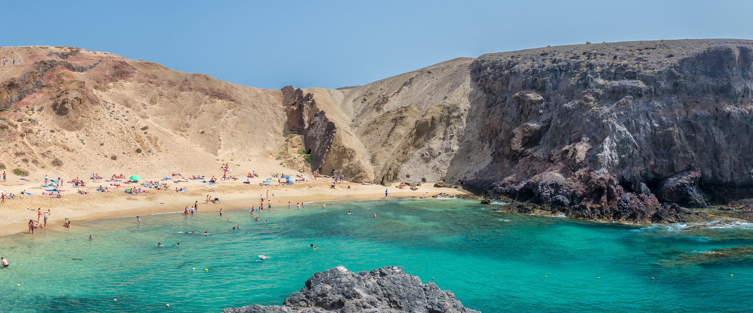 Beach with people swimming and sunbathing, surrounded by rocky cliffs.