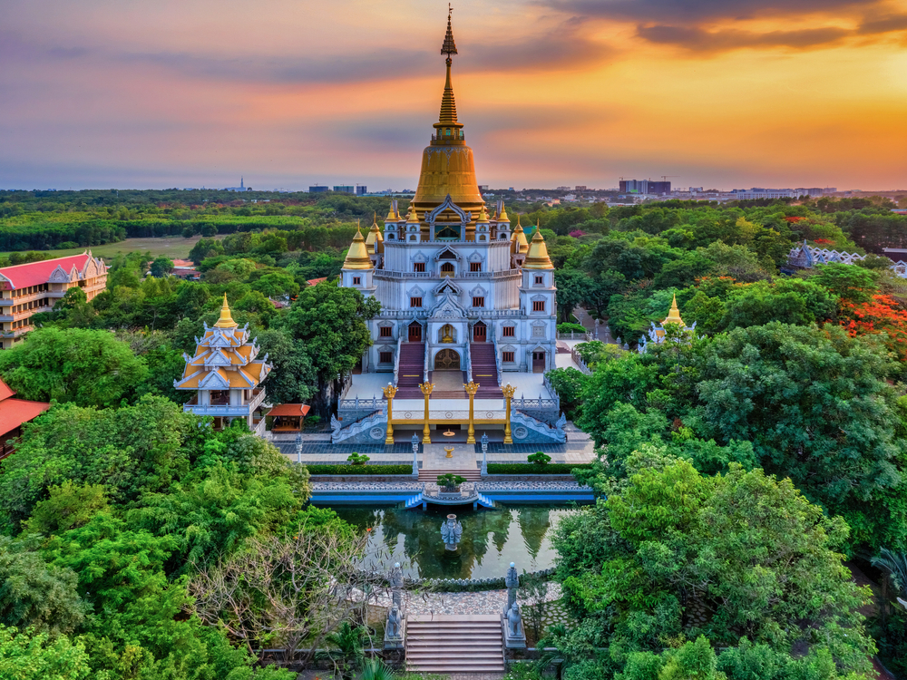 An ornate white and gold temple with a tall spire, surrounded by lush green trees at sunset.