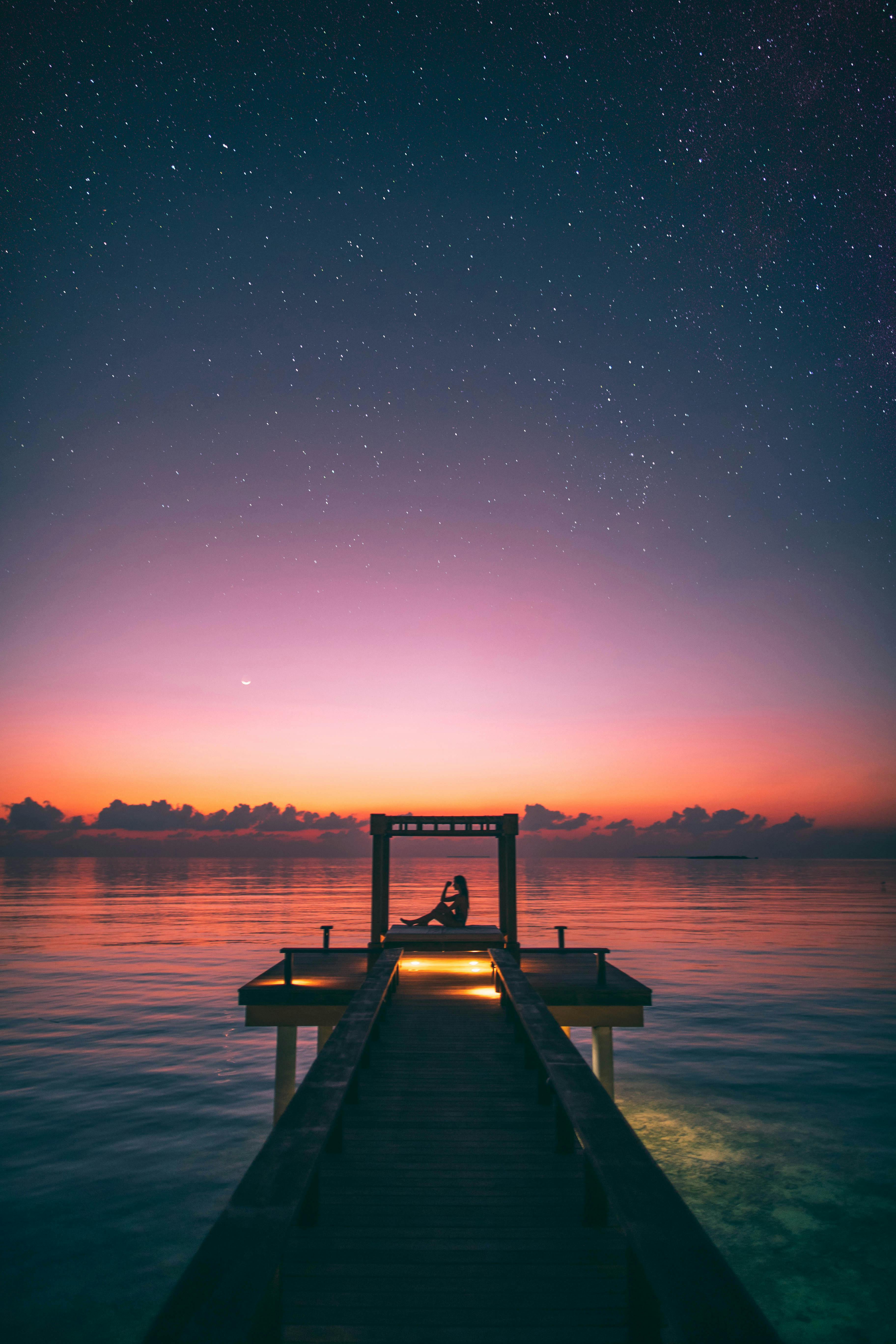 Silhouette of person on pier at sunset with starry sky.