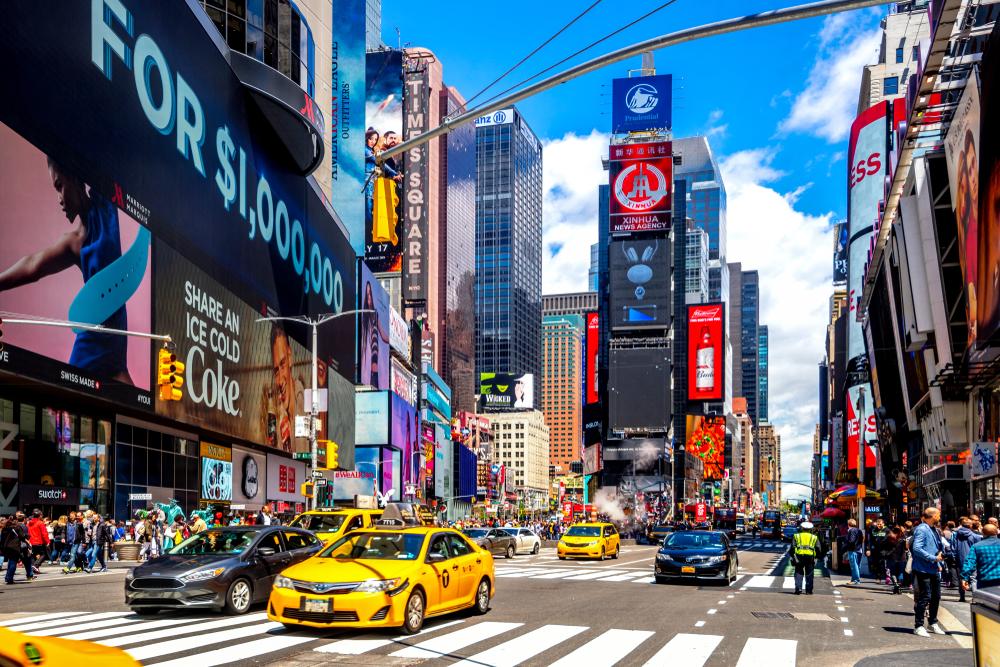 A bustling street view of Times Square on a sunny day, with yellow taxis and large billboards.