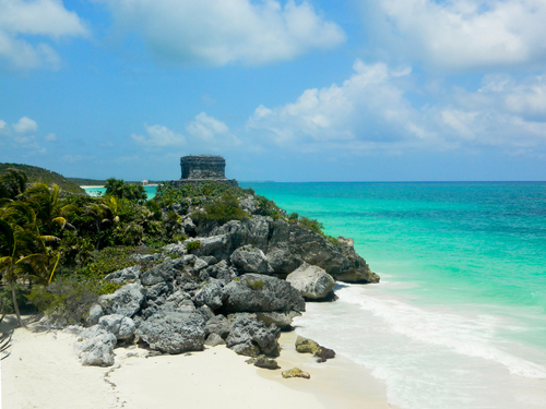 Stone ruins on a rocky cliff overlooking a tropical beach with turquoise water and a partly cloudy sky.