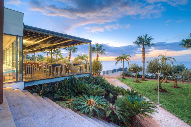 An outdoor restaurant patio with palm trees and landscaped gardens overlooking the ocean at sunset.