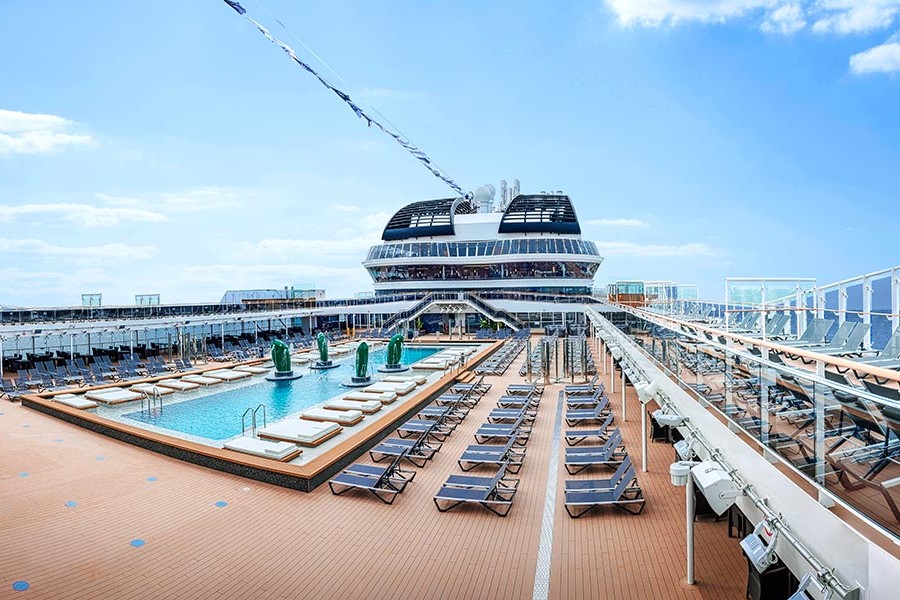 Cruise ship deck with pool, sun loungers, and clear sky.