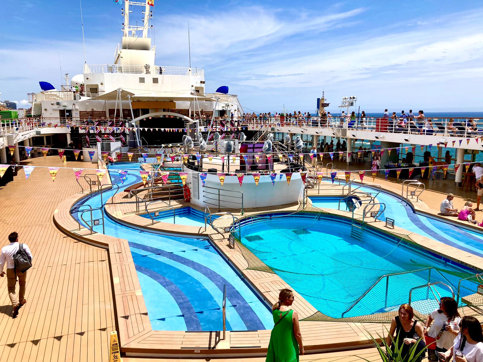 Cruise ship deck with pools, people, and colorful flags under a blue sky.