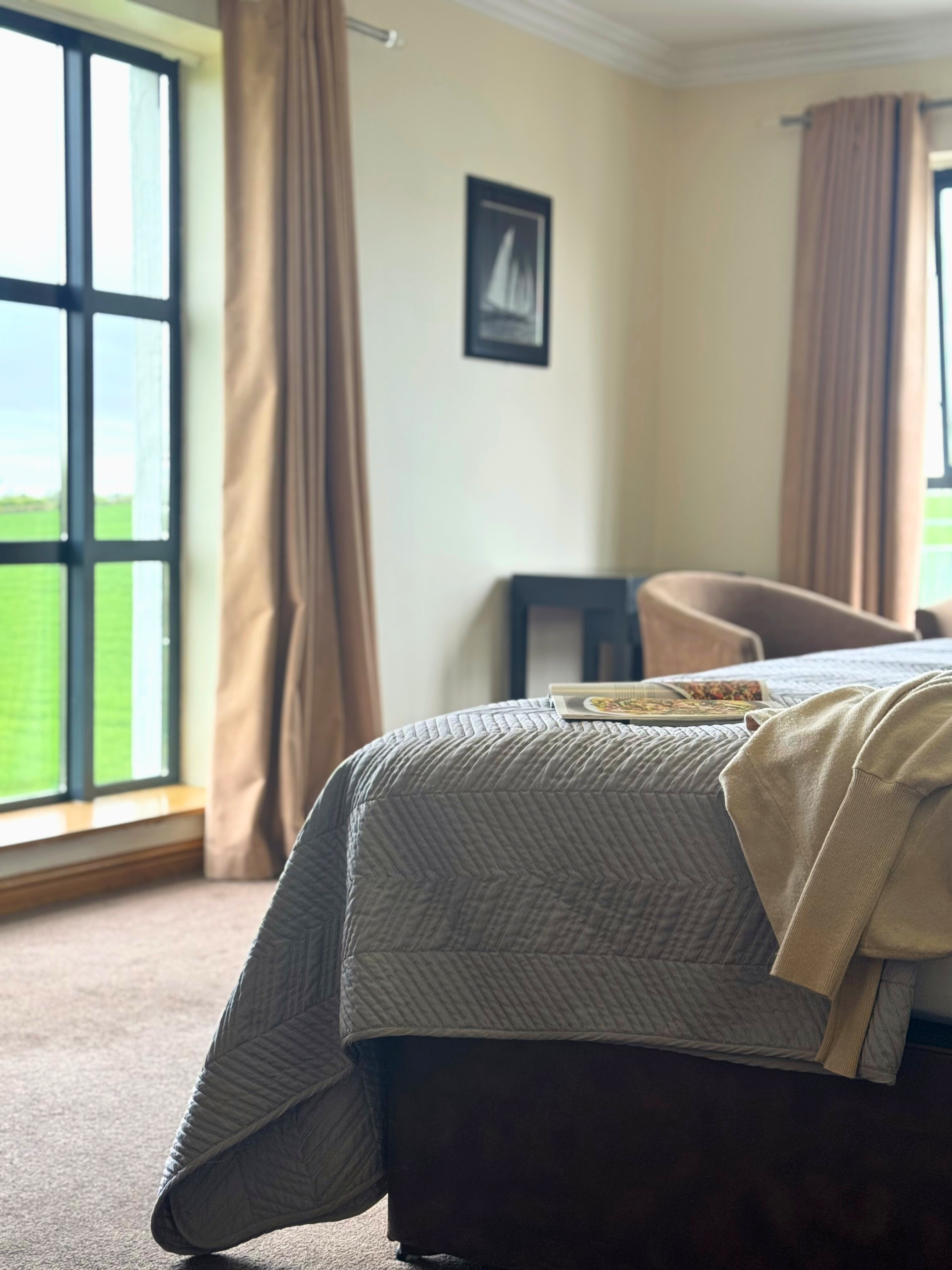 A corner of a neatly made bed in a room with a window overlooking a green field.