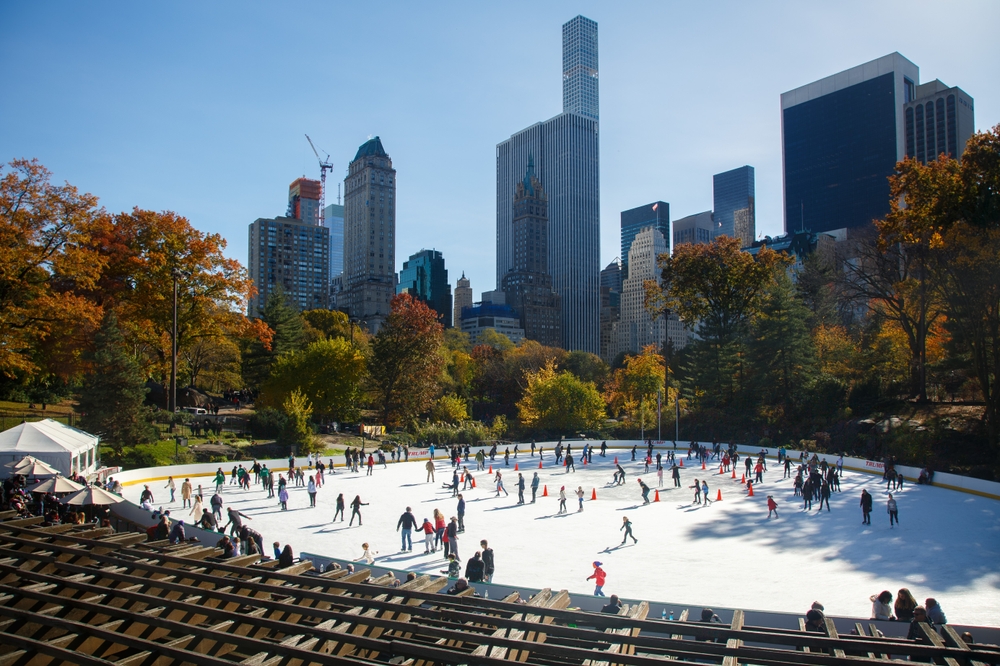 People ice skating at an outdoor rink in a park with a city skyline in the background.