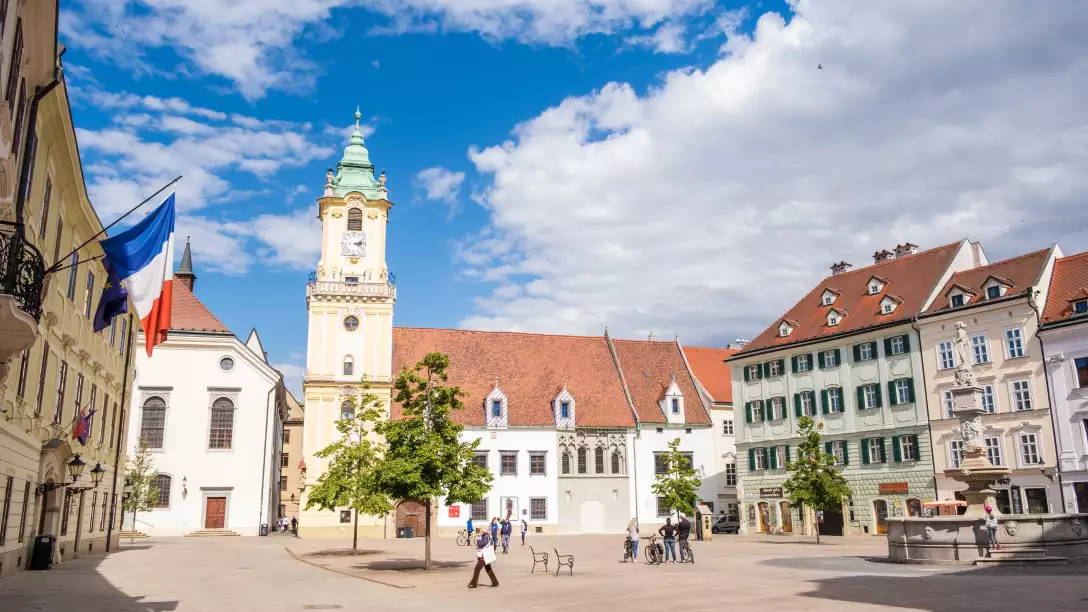 A sunny European town square with a clock tower, historic buildings, and people walking around.