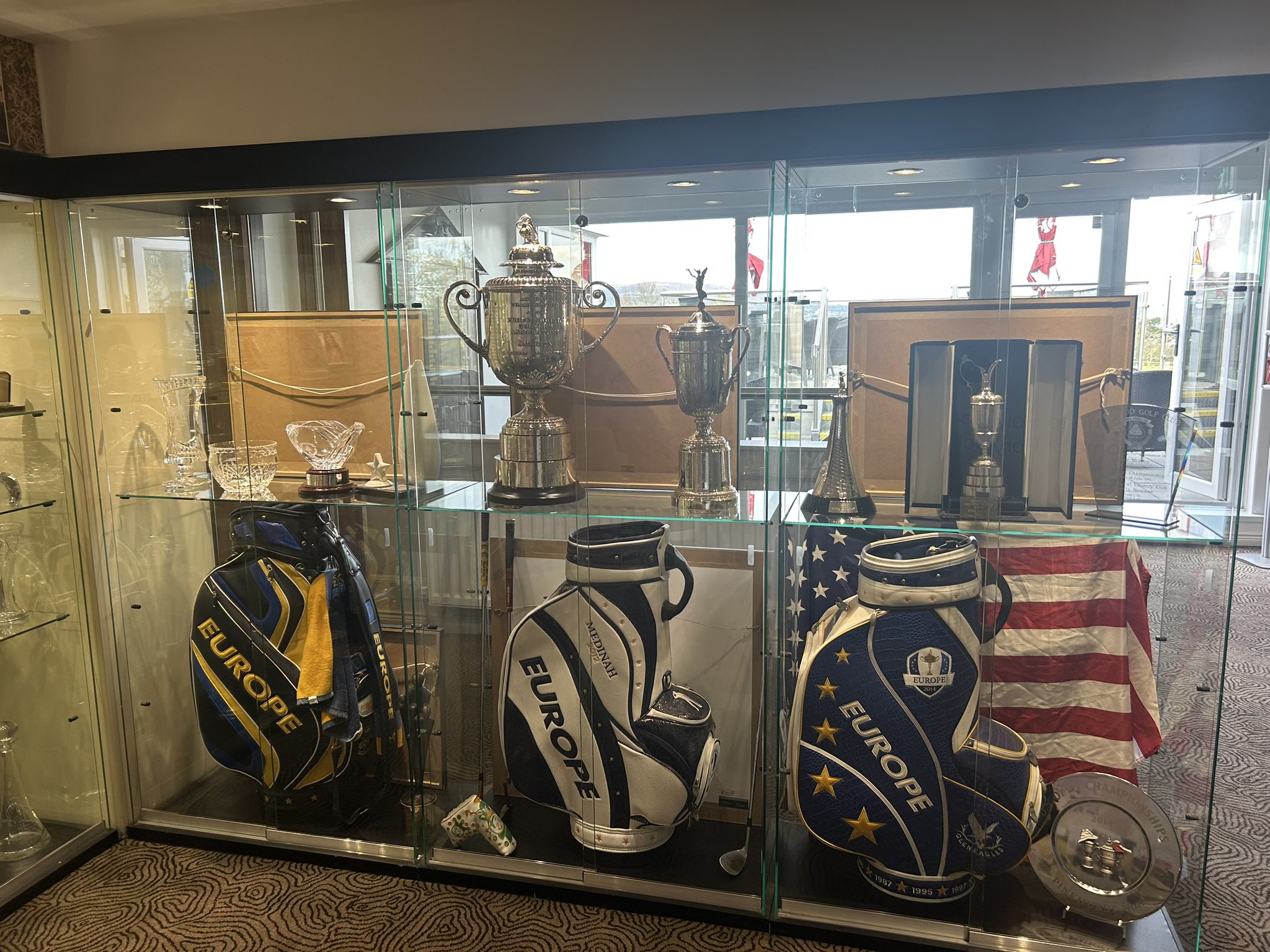 Glass display case with golf trophies, bags, and an American flag.