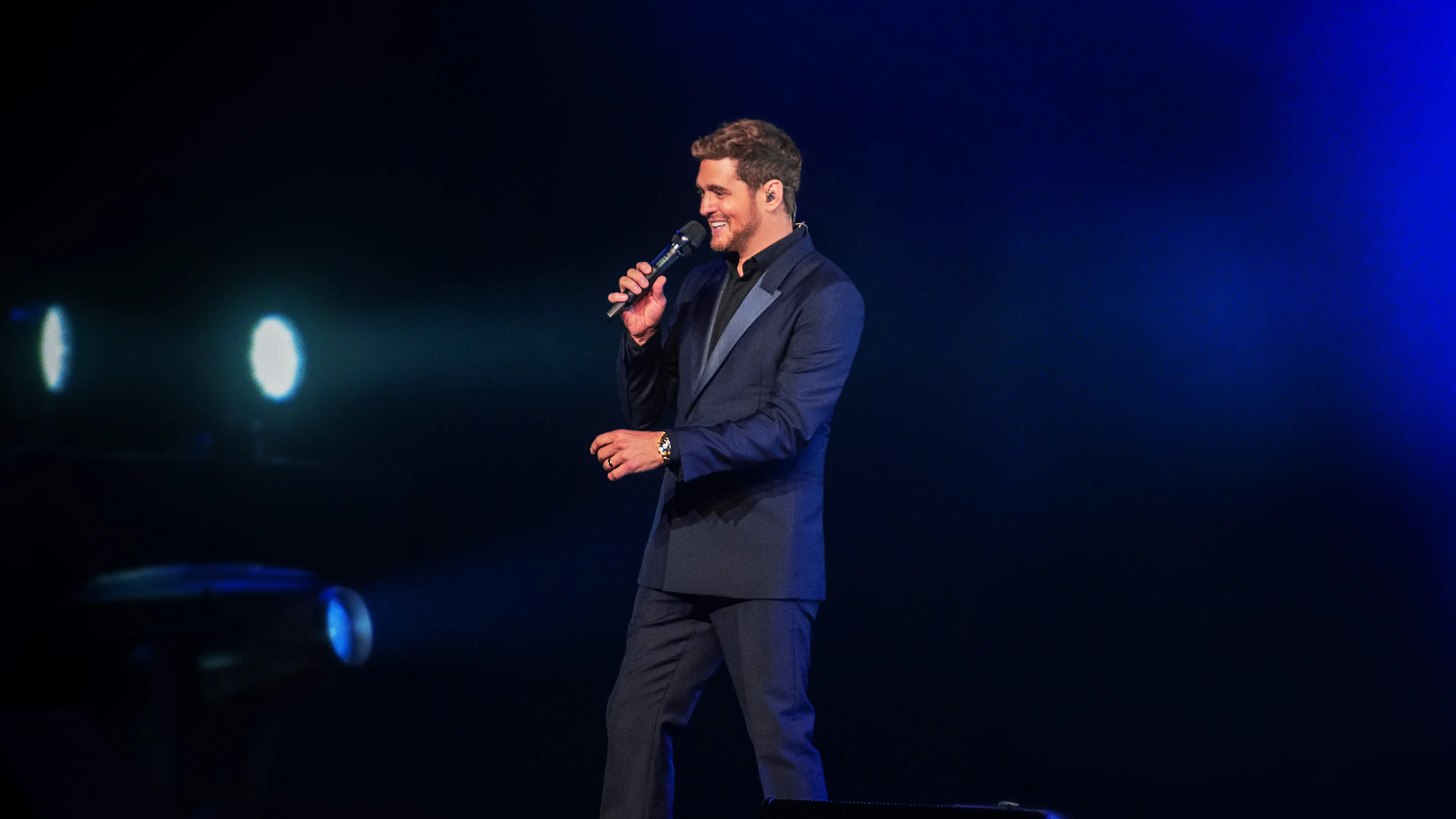 A man in a dark blue suit sings into a microphone on a dark stage with blue lights.