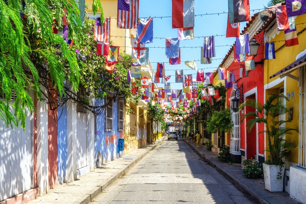 A narrow city street with colorful buildings and international flags strung overhead on a sunny day.