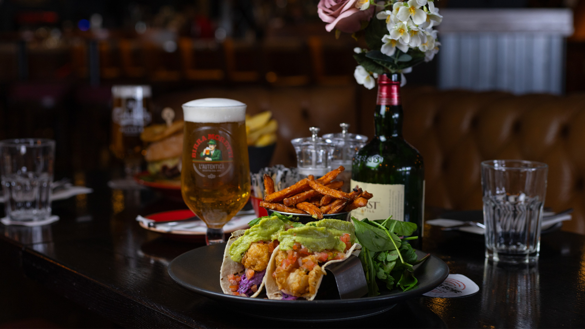 A plate of tacos with sweet potato fries and a glass of beer on a restaurant table.