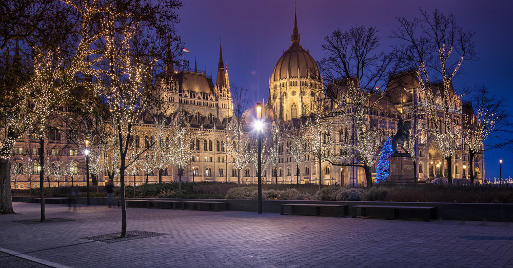 A large, ornate building at dusk, with trees wrapped in white lights in the foreground plaza.