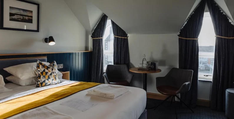 A hotel room with a double bed and a seating area with two chairs next to two windows.