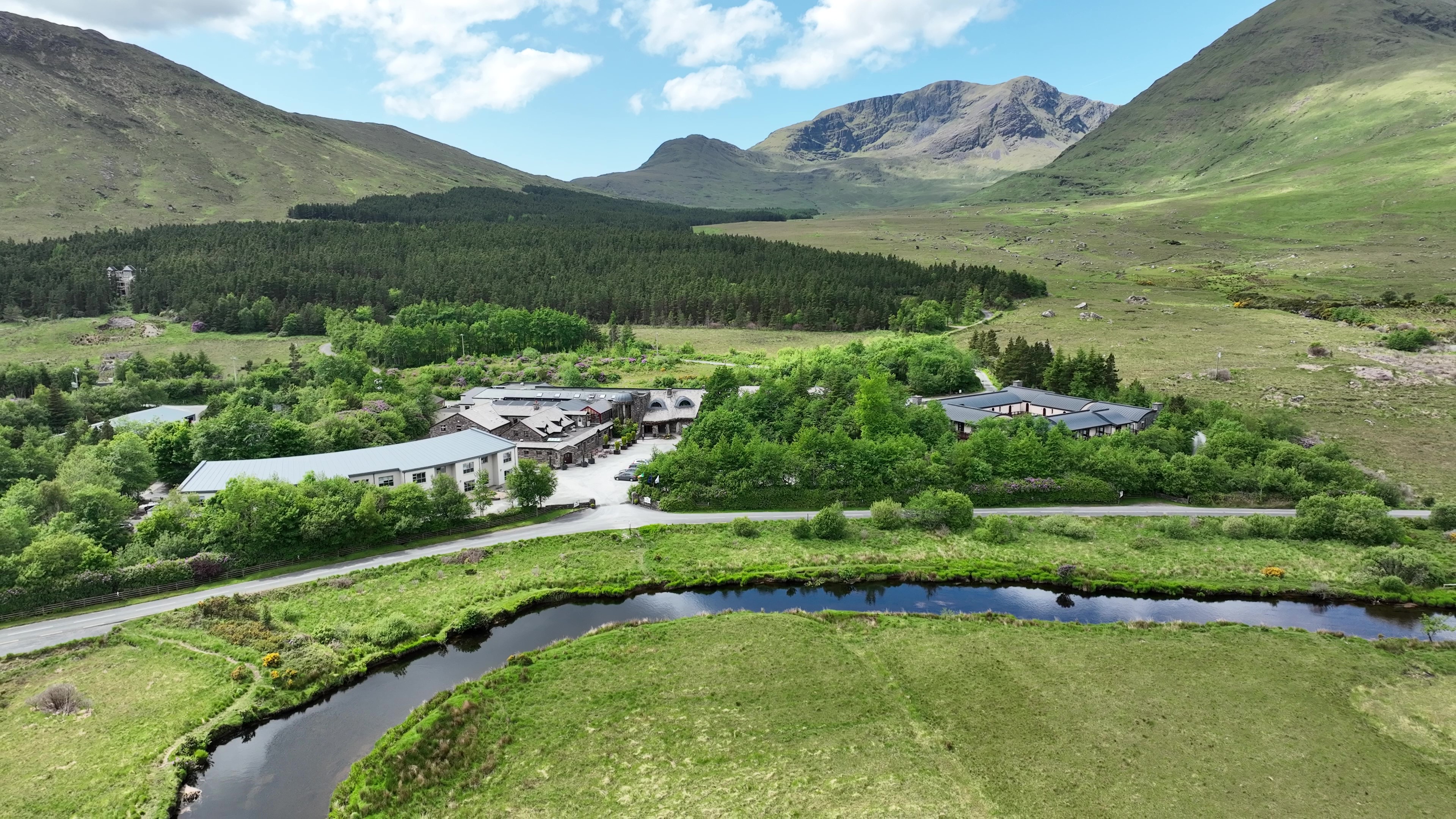 An aerial view of a hotel complex in a lush green valley surrounded by mountains and a river.
