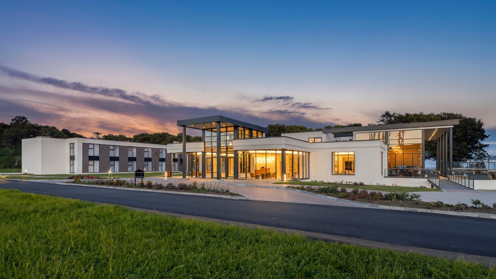 Modern building with large windows at sunset, surrounded by greenery.
