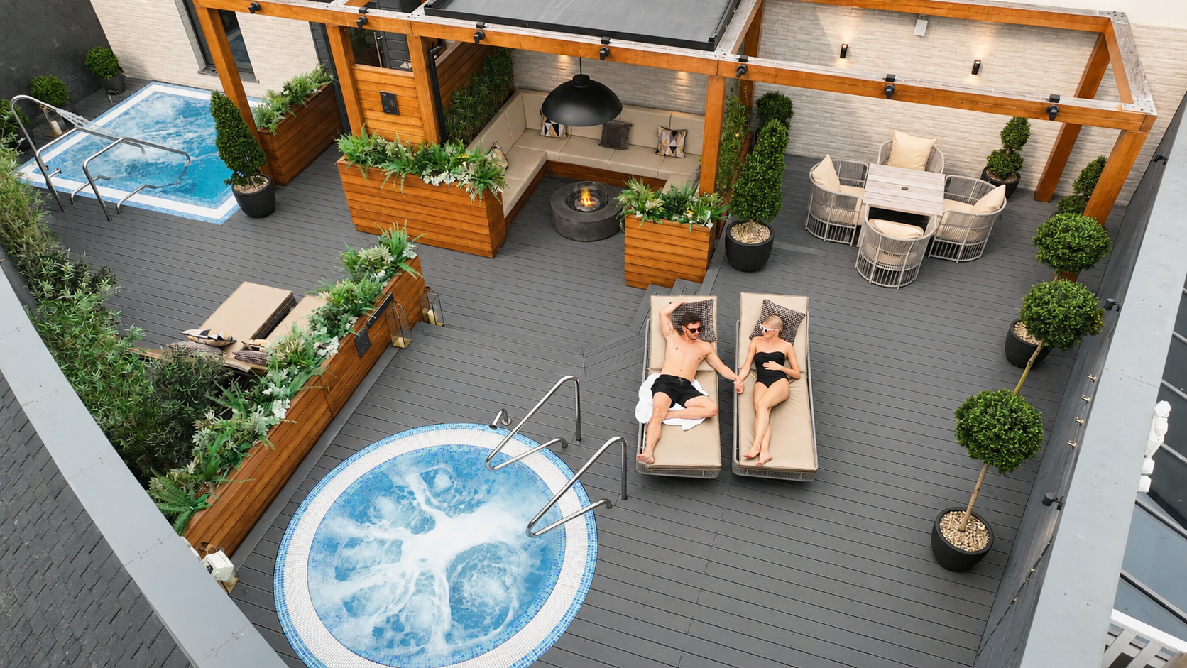 An overhead view of a couple sunbathing on a luxurious rooftop patio with two hot tubs and lounge furniture.