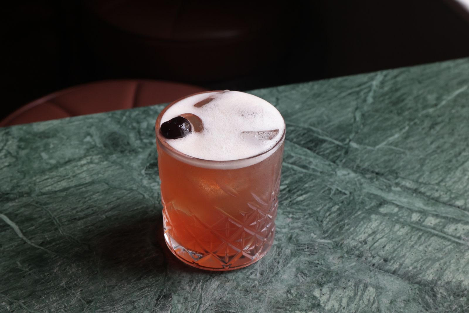 An amber-colored cocktail with a frothy top and cherry garnish sits on a green marble countertop.