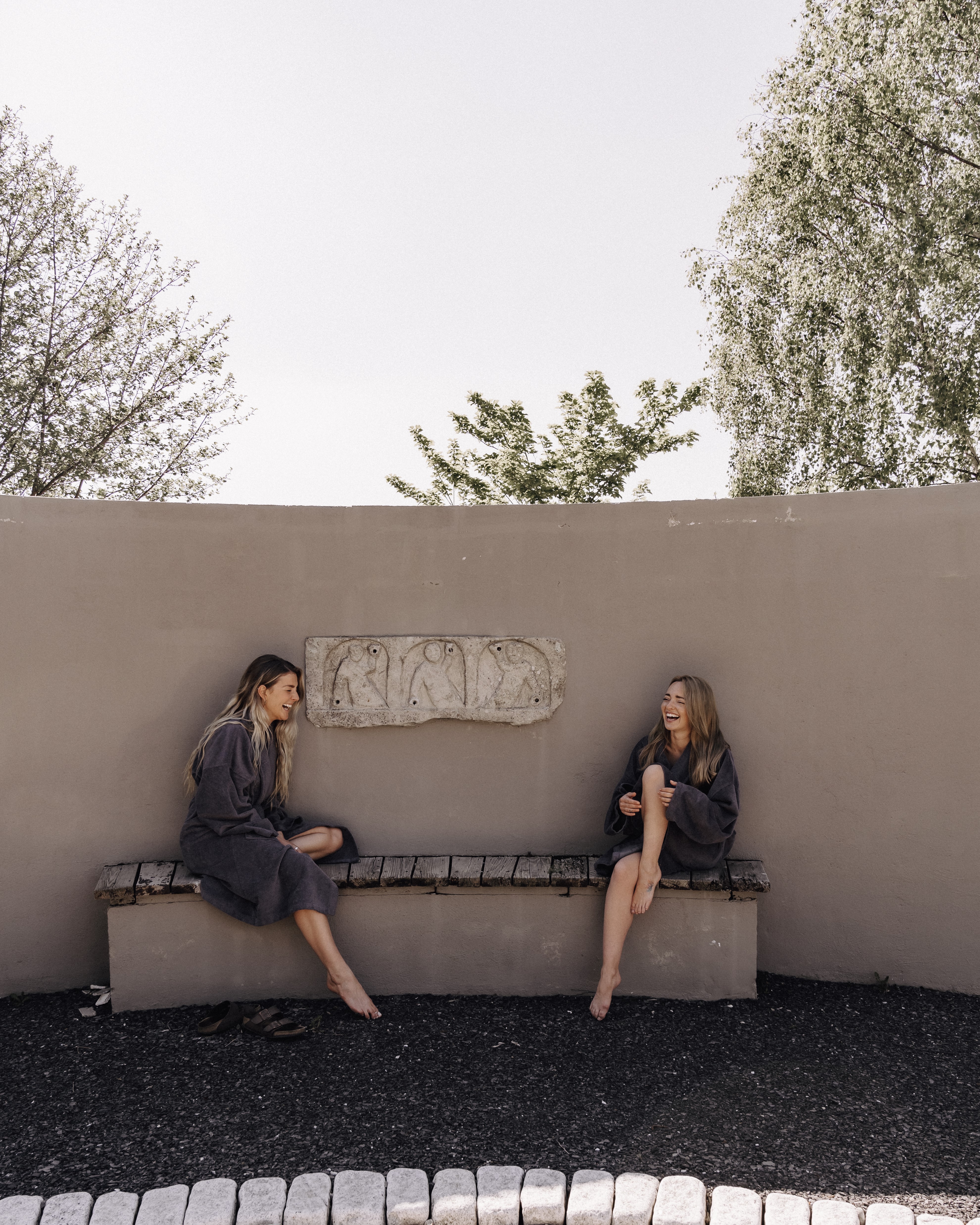 Two women in robes sitting on a bench, laughing, with trees in the background.