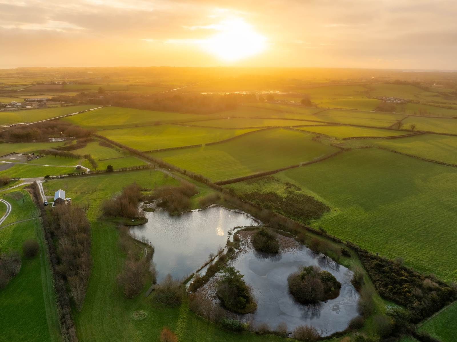 Aerial view of a lake and green fields at sunset, with the sun casting a golden glow.