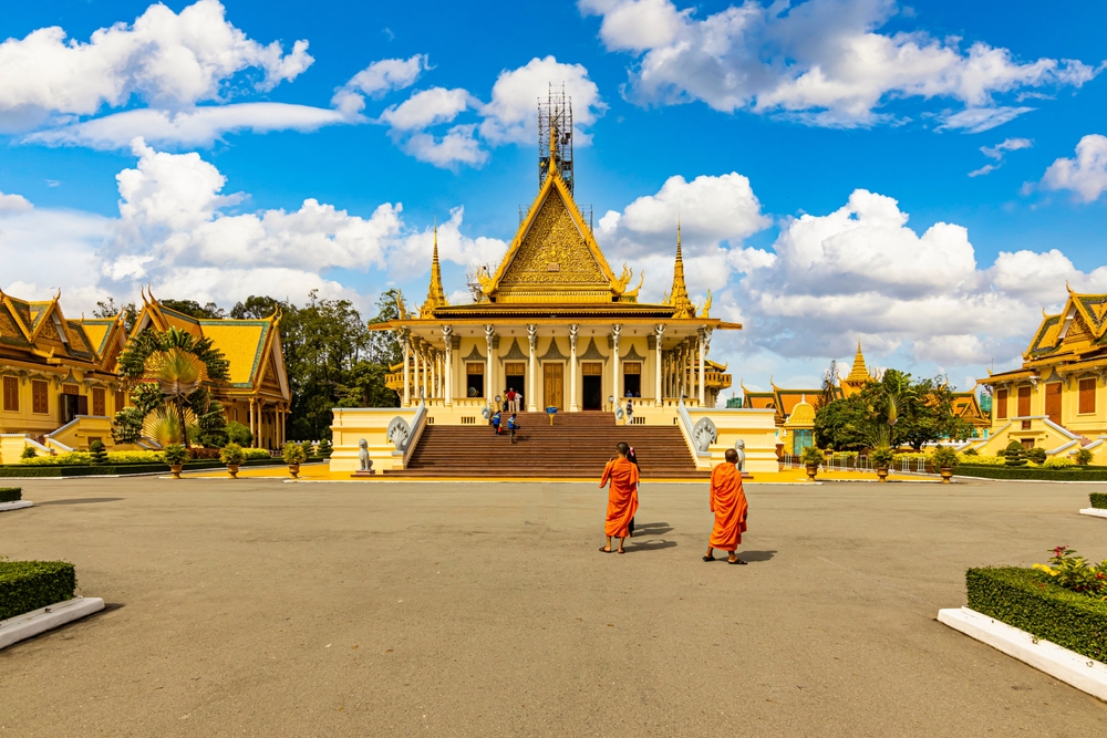 Two monks in orange robes walk across a large courtyard towards an ornate temple with a golden roof.