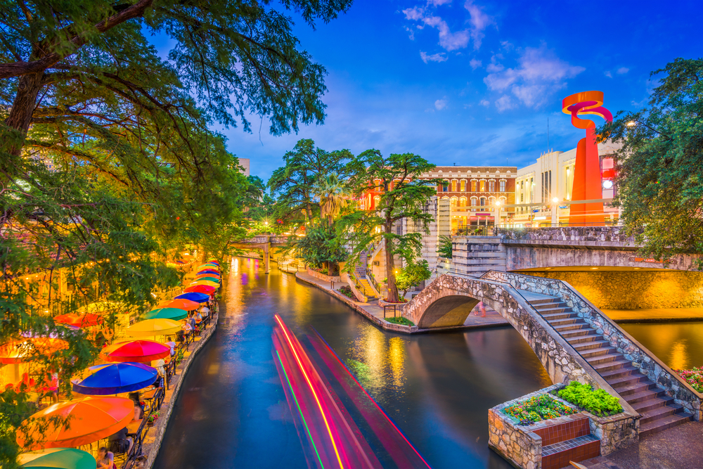 A city river walk at dusk with colorful umbrellas, a stone bridge, and light trails from a boat.