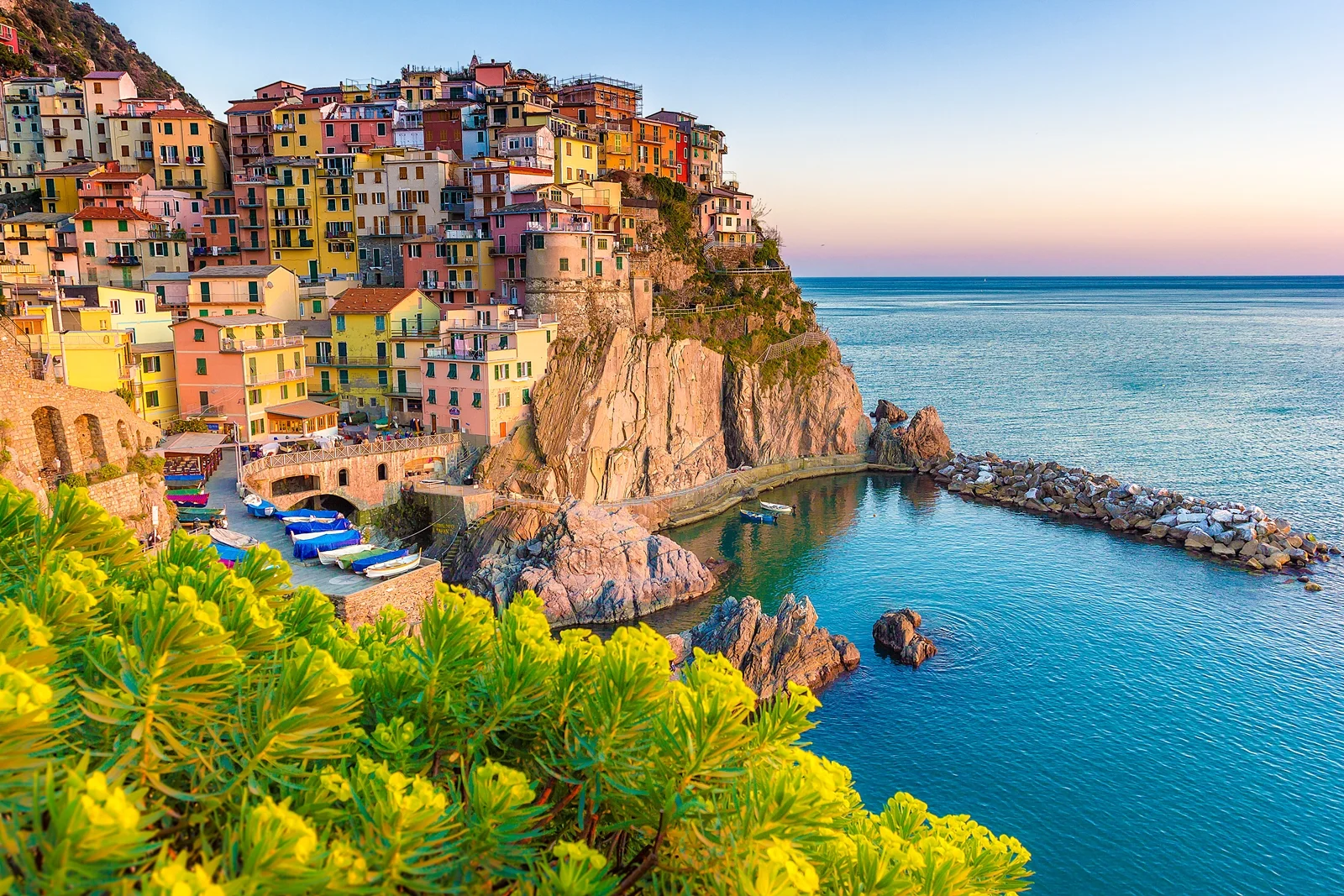 Colorful buildings clustered on a rocky cliff overlooking a tranquil sea at dusk, with boats in a cove.