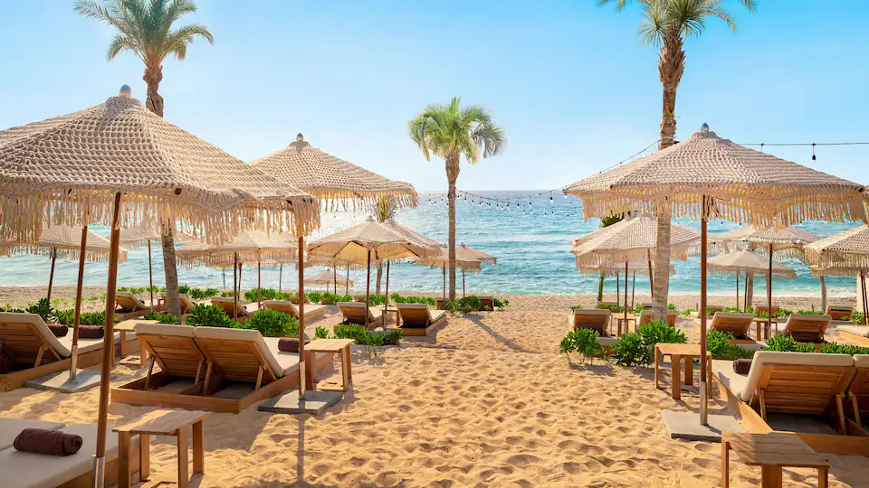 Lounge chairs and woven umbrellas with fringe on a sandy beach next to the ocean and palm trees.