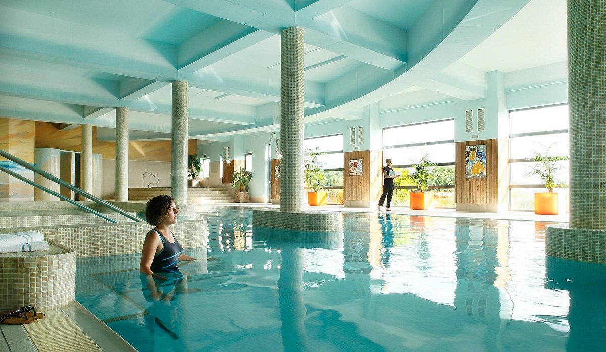 Indoor pool with a person swimming and another standing by large windows.