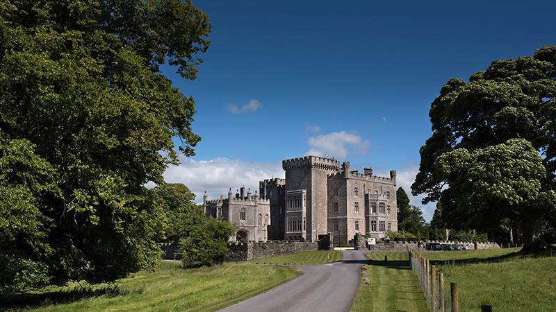 A large stone castle at the end of a road, framed by green trees under a blue sky.
