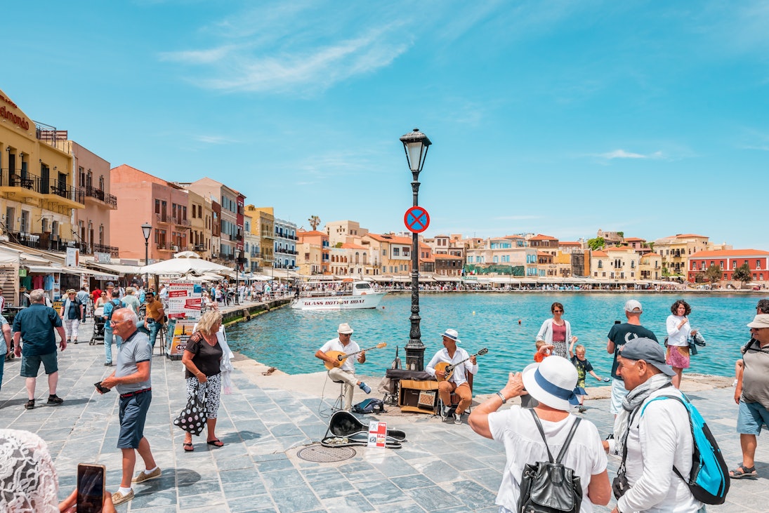 Street musicians perform by a waterfront with colorful buildings and tourists.