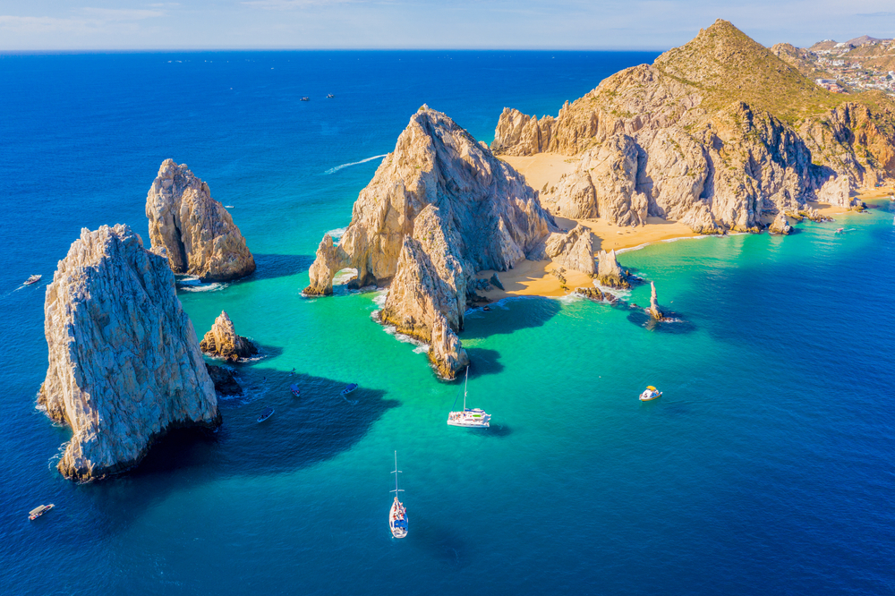 An aerial view of large rock formations and a natural arch in turquoise ocean waters with several boats.